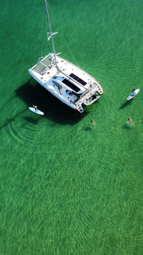 Aerial view of a catamaran on clear green water with two paddleboarders nearby.
