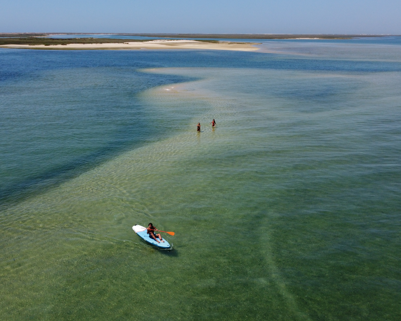 Person kayaking in clear water near shallow sandbar with two people, distant land visible.