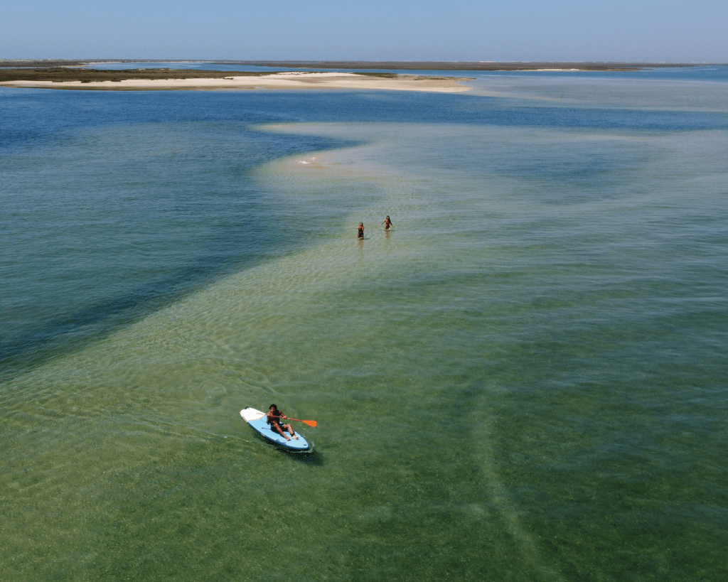Person kayaking in clear water near shallow sandbar with two people, distant land visible.