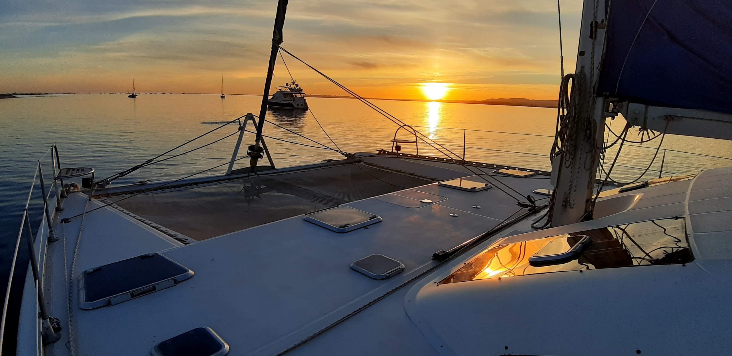 Sailboat deck at sunset with calm water and distant boats.