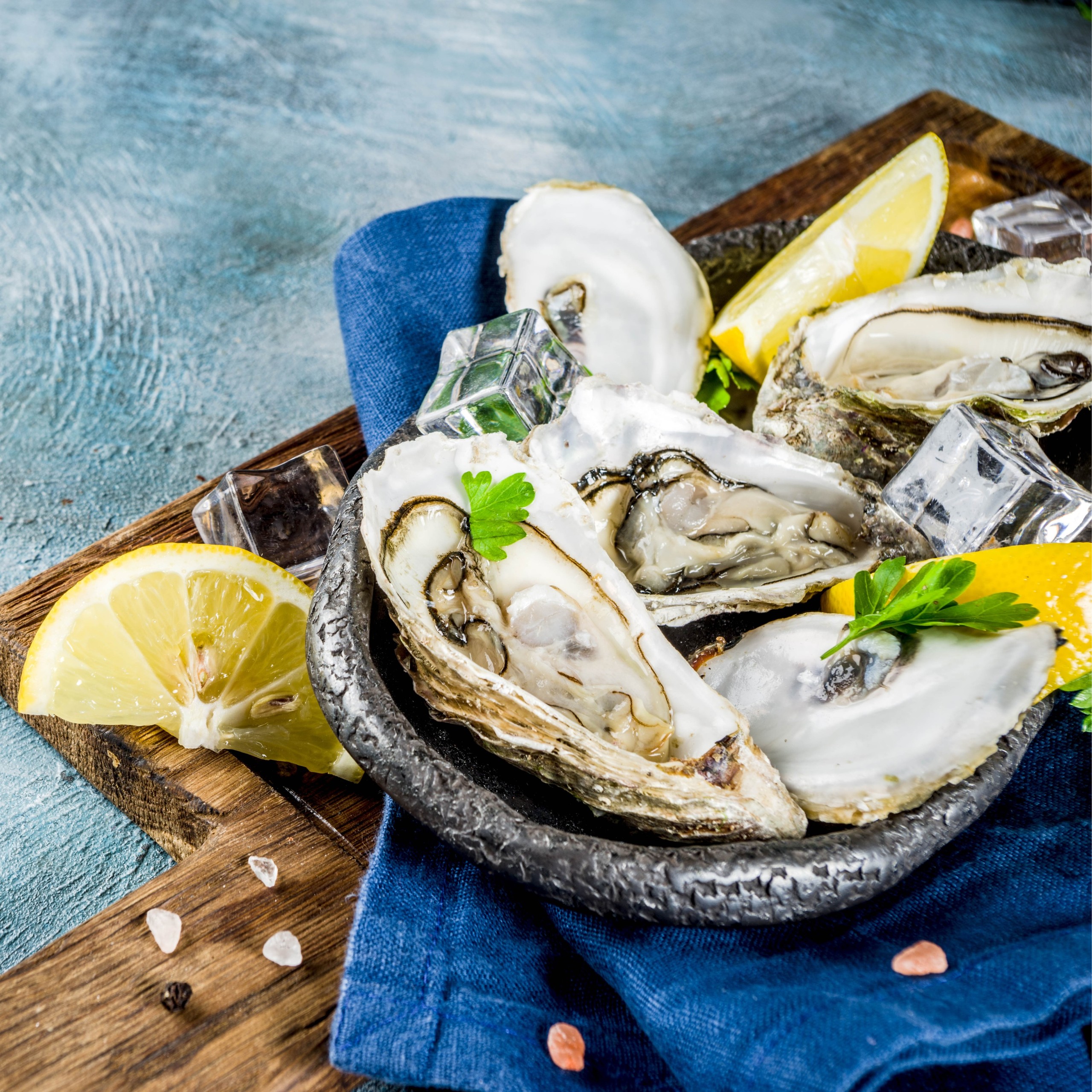 Oysters on a plate with lemon wedges, parsley, ice cubes, and salt on a blue cloth.