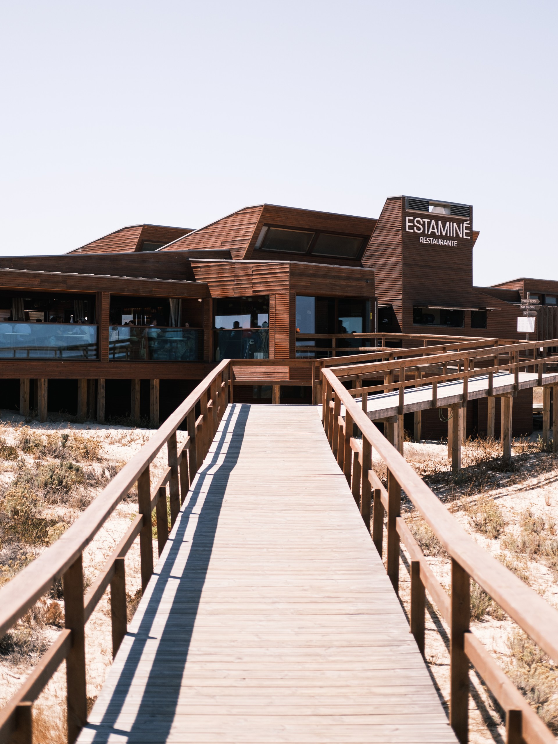 Wooden walkway leading to a modern beachside restaurant with large windows.