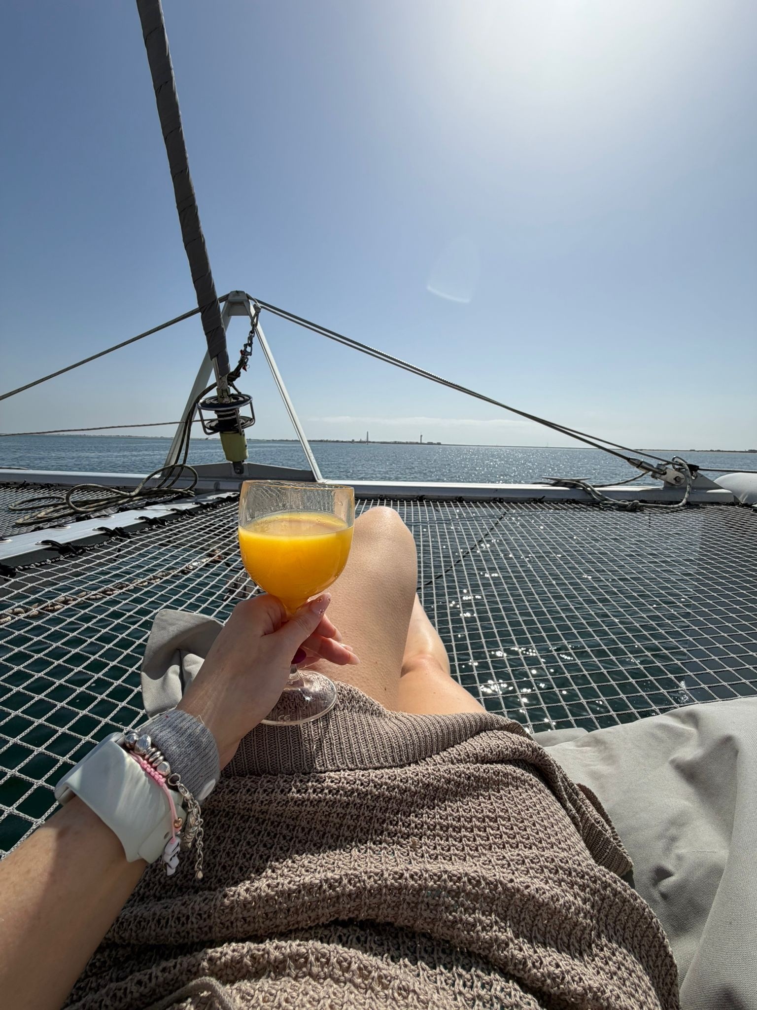Person relaxing on a boat net with a drink, ocean view.