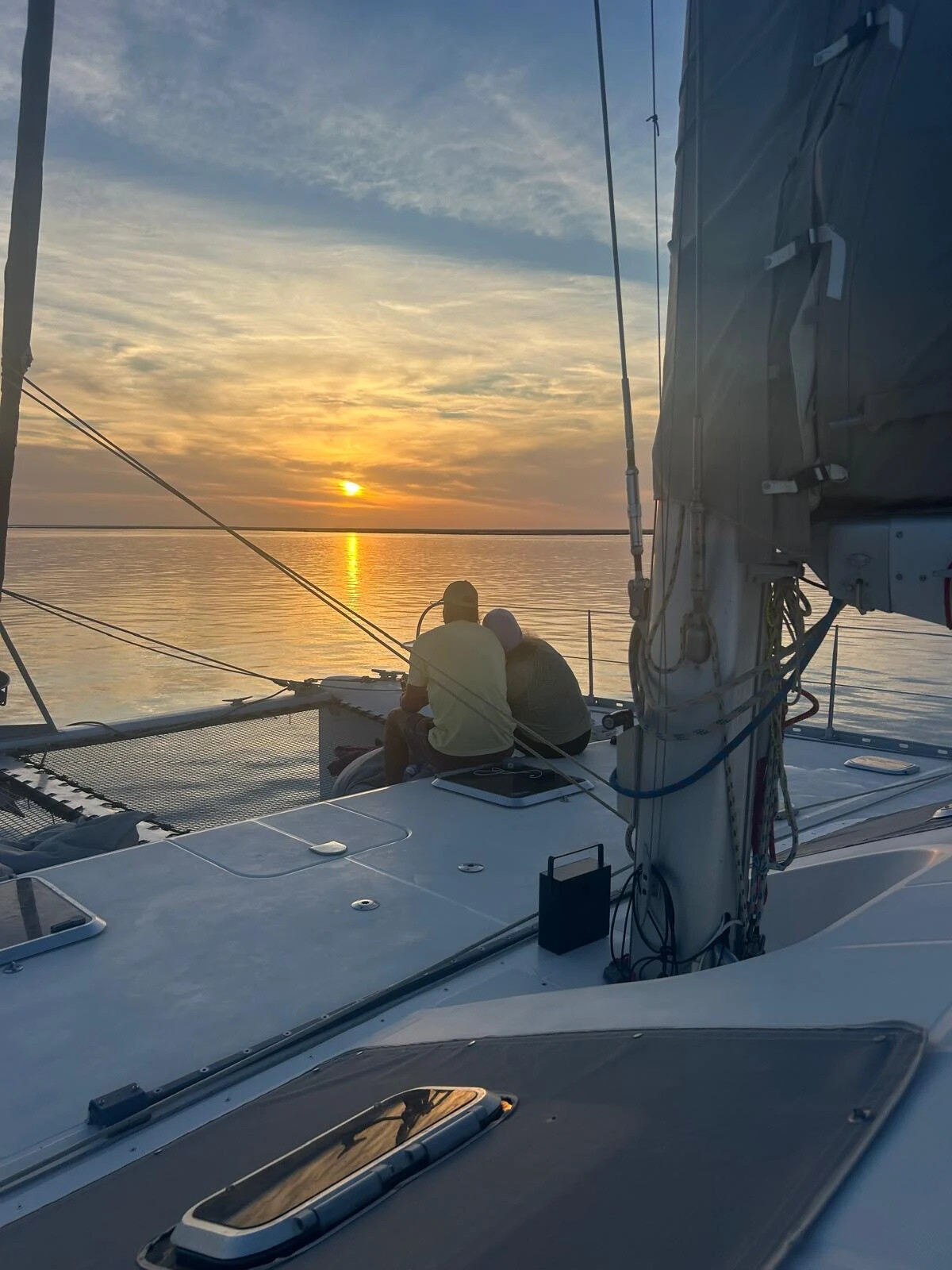 Two people sit on a catamaran deck at sunset over calm water.