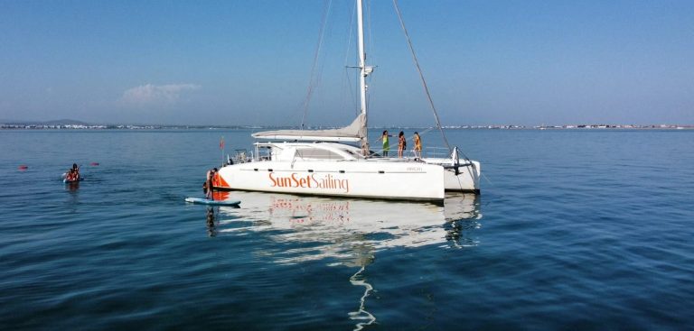 Catamaran named SunSet Sailing on calm water with people on deck and kayaker nearby.