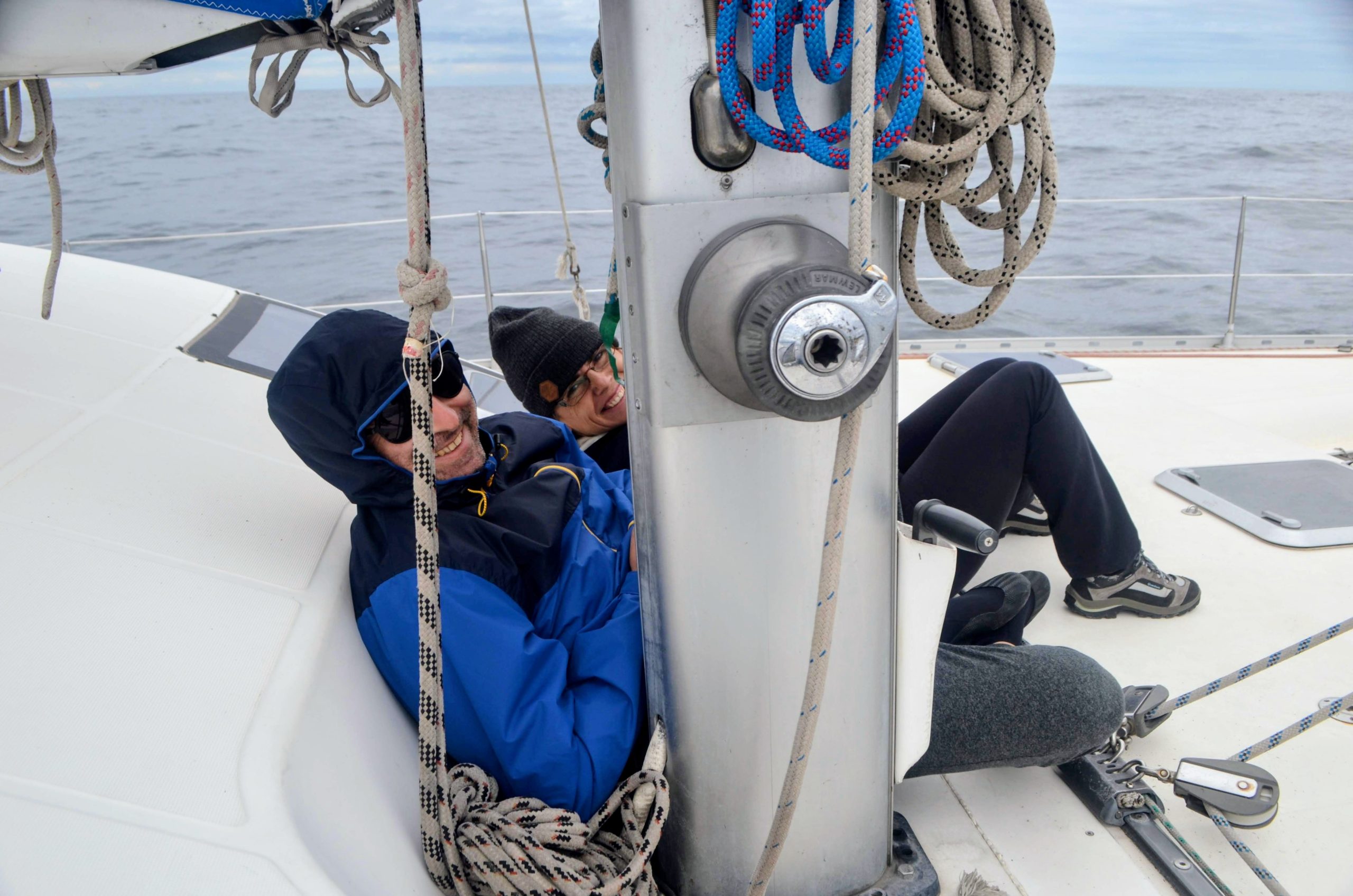 Two people in jackets relax on a sailboat, leaning against the mast with ropes and winches visible.
