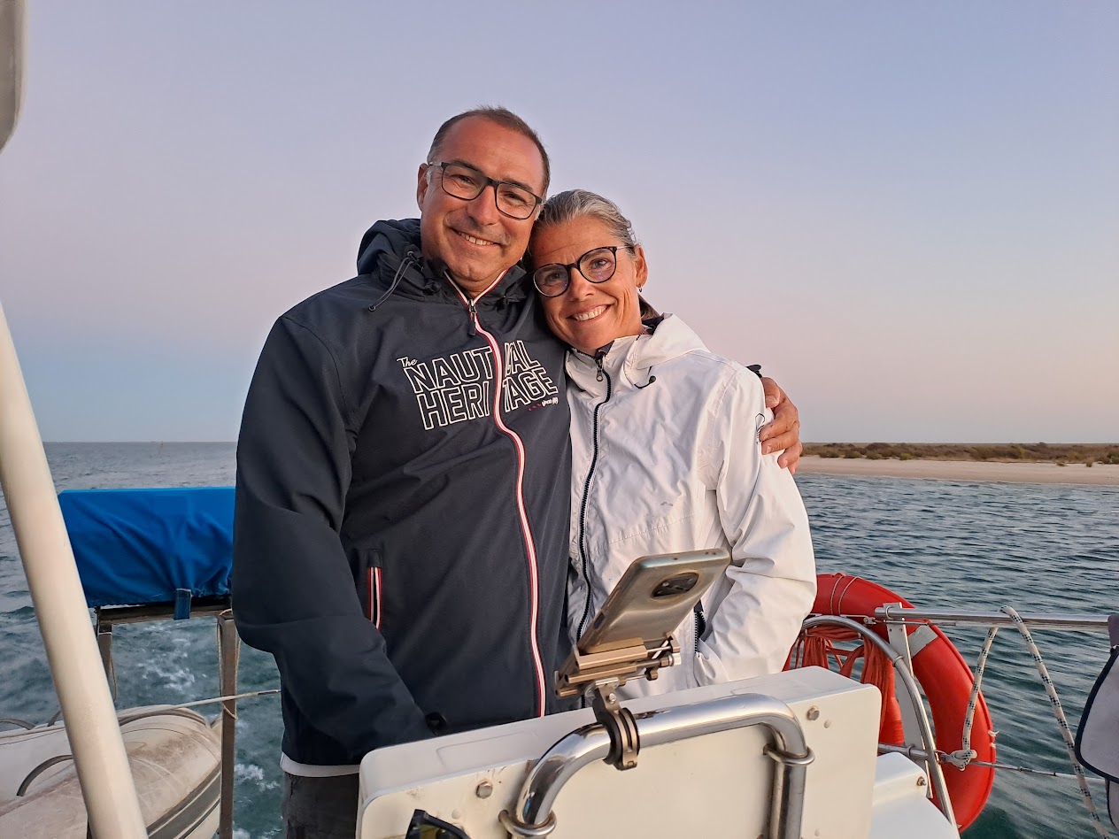 Two people smiling on a boat, one wearing a black jacket and the other in white, with ocean in the background.