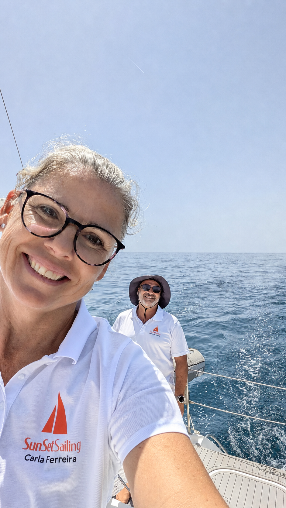 Two people smiling on a sailboat, wearing white shirts with ocean in the background.