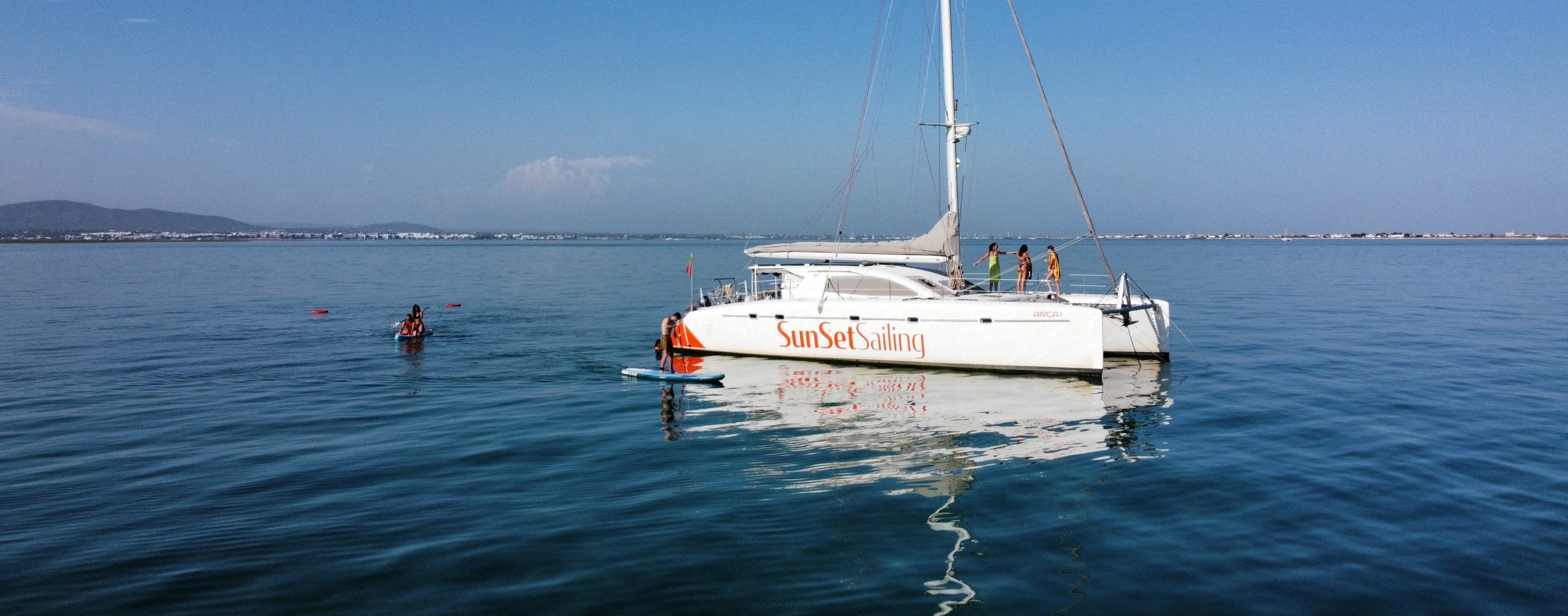 Catamaran sailing on calm water with clear sky and distant shoreline.