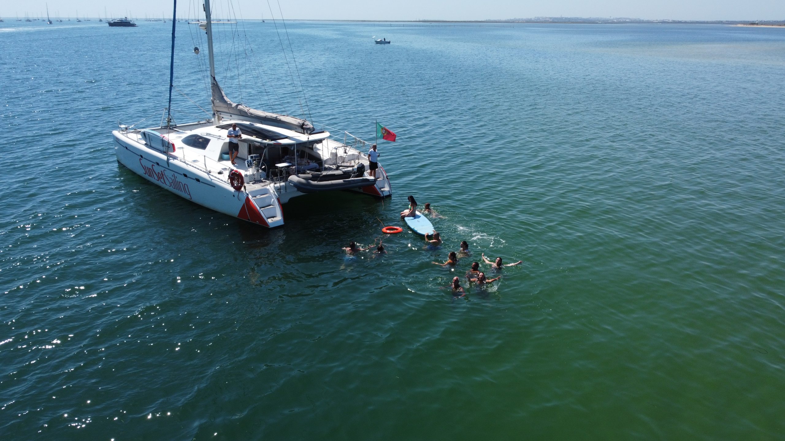 Catamaran in ocean with people swimming nearby on a sunny day.